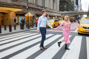 Couple holding hands crossing a zebra crosswalk near Grand Central Terminal with yellow NYC taxis behind.