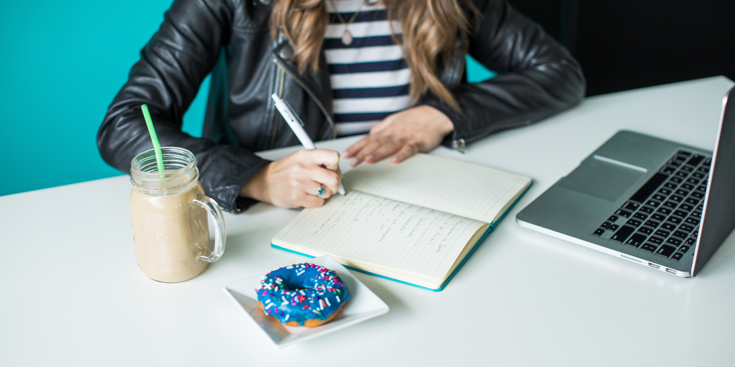 Workspace scene with laptop, notebook, coffee and blue donut on white desk.