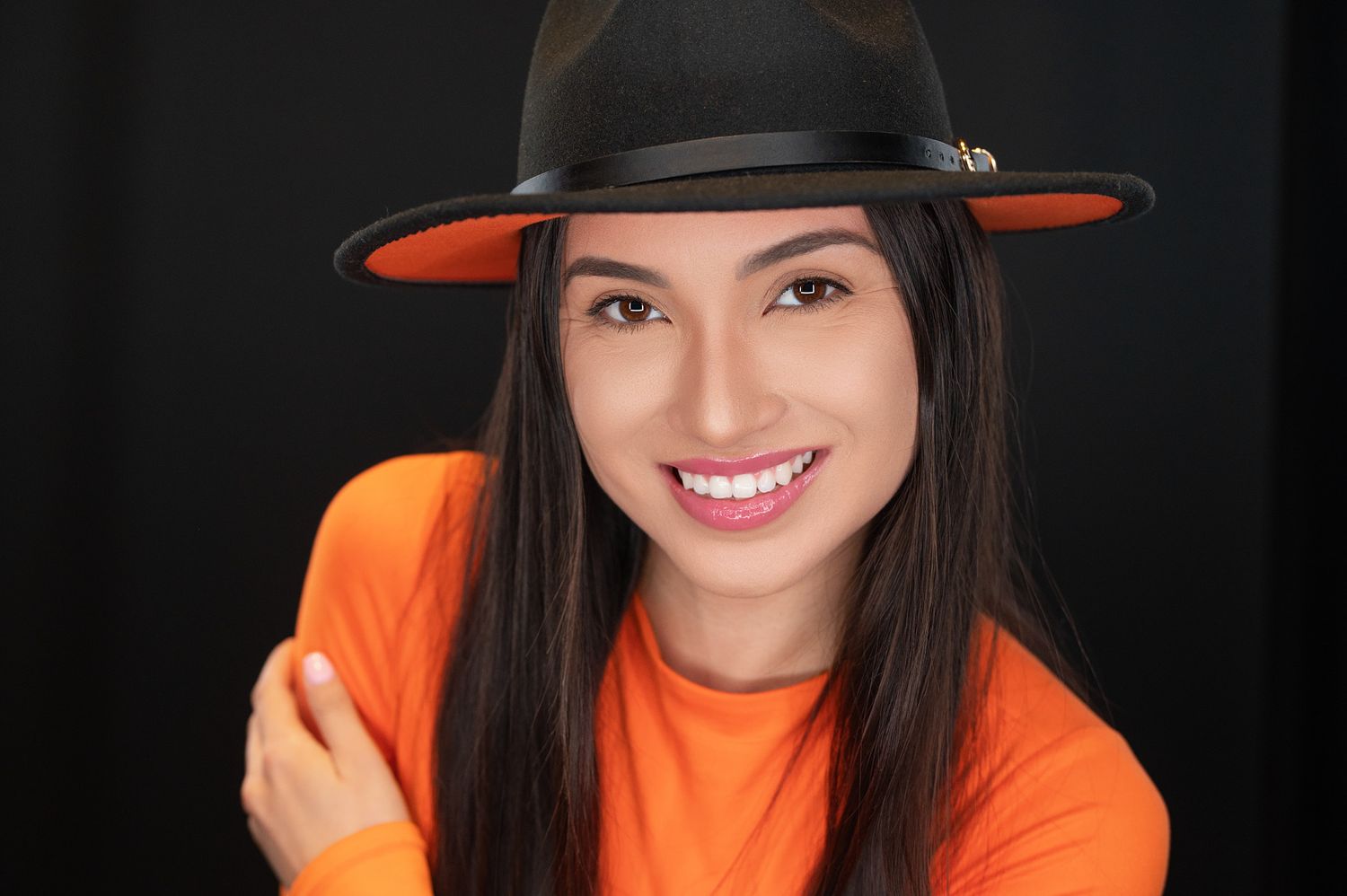 Smiling young woman in orange shirt wearing a black hat against a dark background.