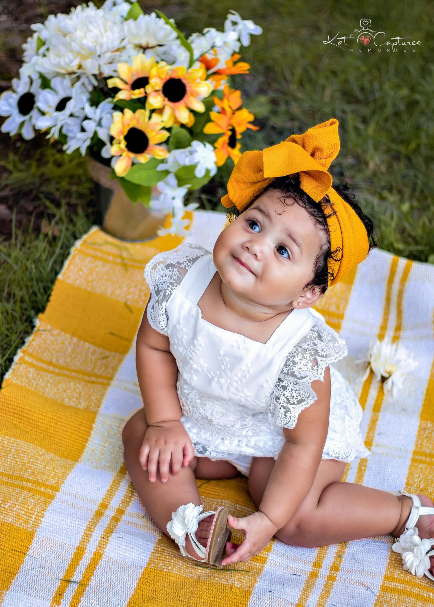 Baby in white lace dress and yellow bow poses on yellow striped blanket with sunflowers.