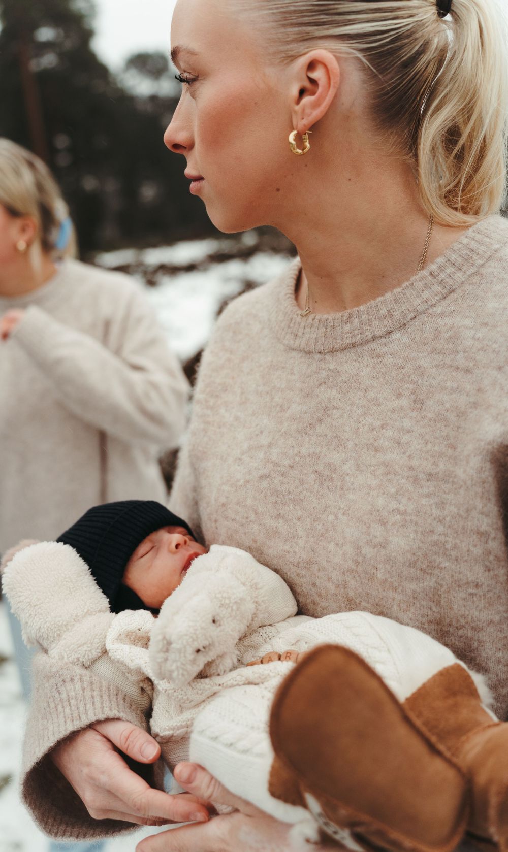 A person in a beige sweater holding a sleeping infant bundled in white winter clothing.