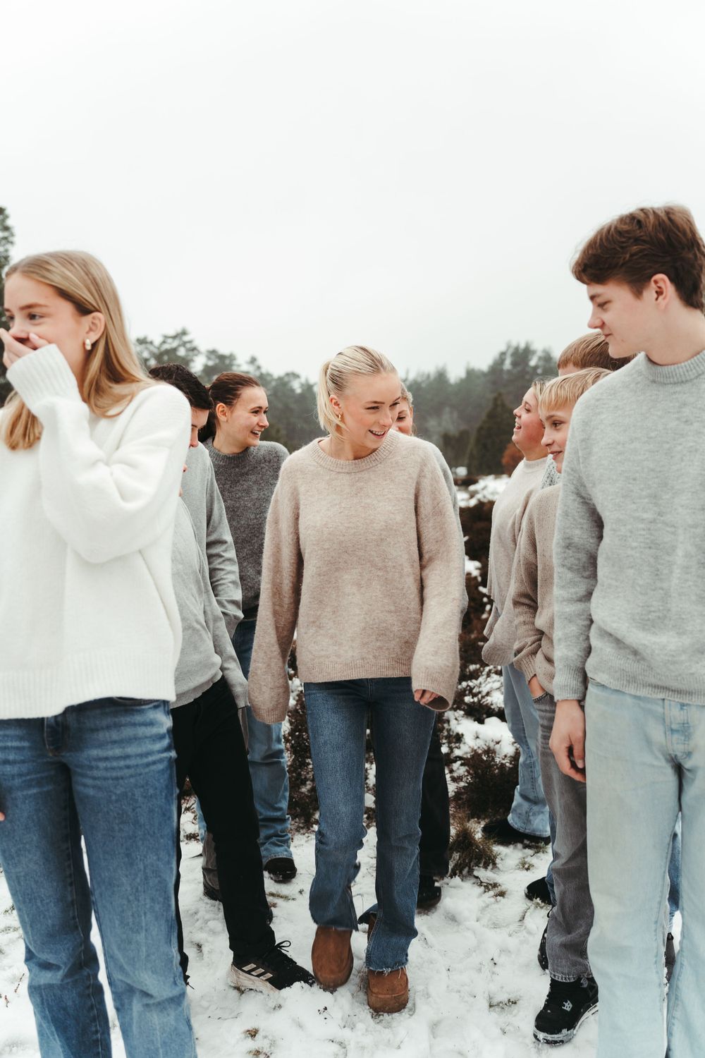 A group in cozy sweaters and jeans standing together on snowy ground with trees in the background.