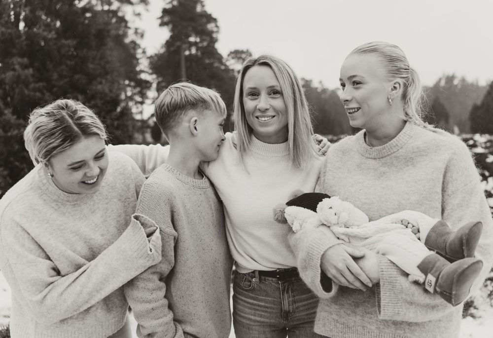 Friends in matching sweaters share a candid moment while holding an infant outdoors.