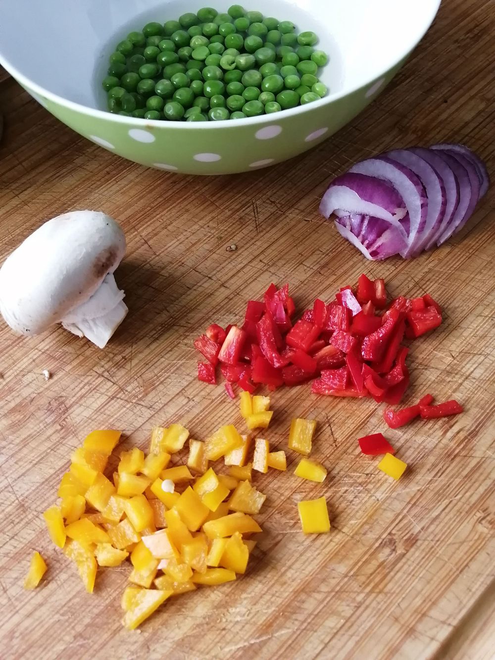 Bread board with a green polka dot bowl with fresh peas, some red onion,  red and yellow pepper and a mushroom
