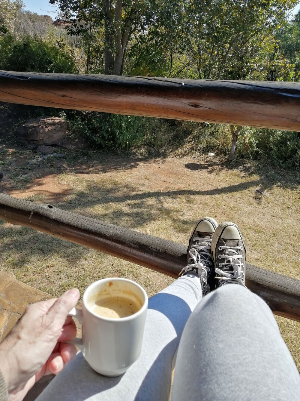 View of women's legs wearing jeans and black allstars holding a cup of coffee in the early morning sun