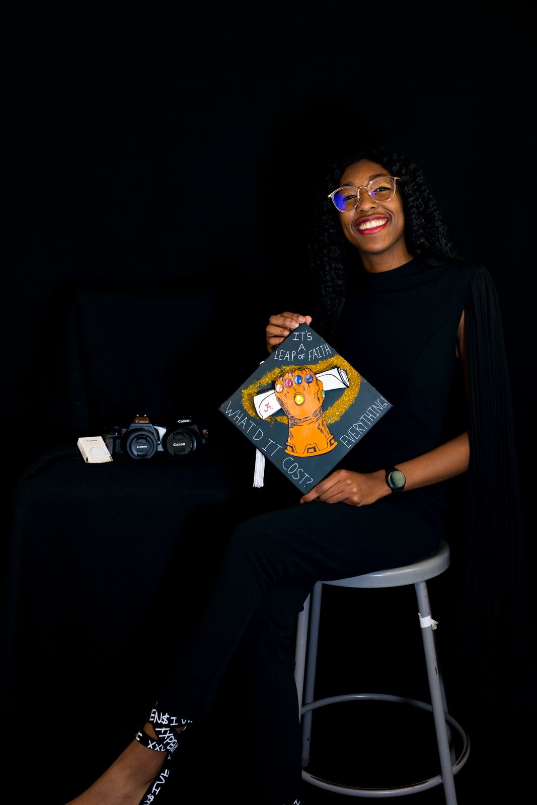 Kassidy/KLo sitting on a stool holding a grad cap and three cameras next to them on a table.
