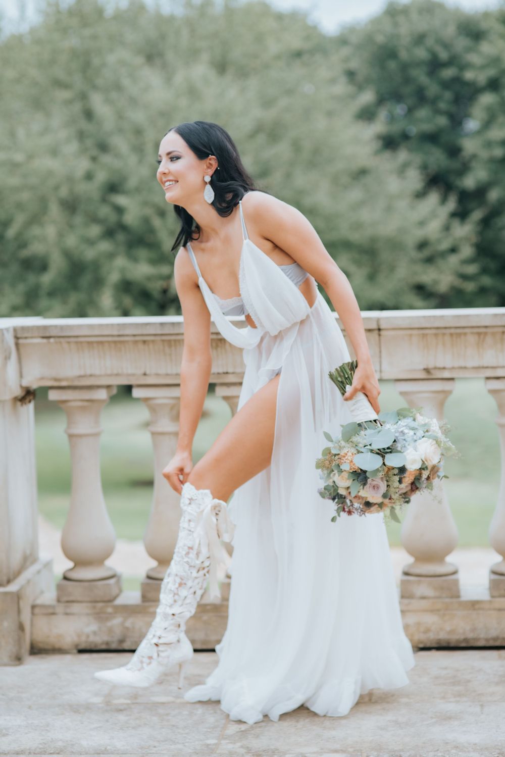 Ethereal bridal portrait showcasing white flowing dress and vintage boots on elegant stone balcony with floral bouquet.