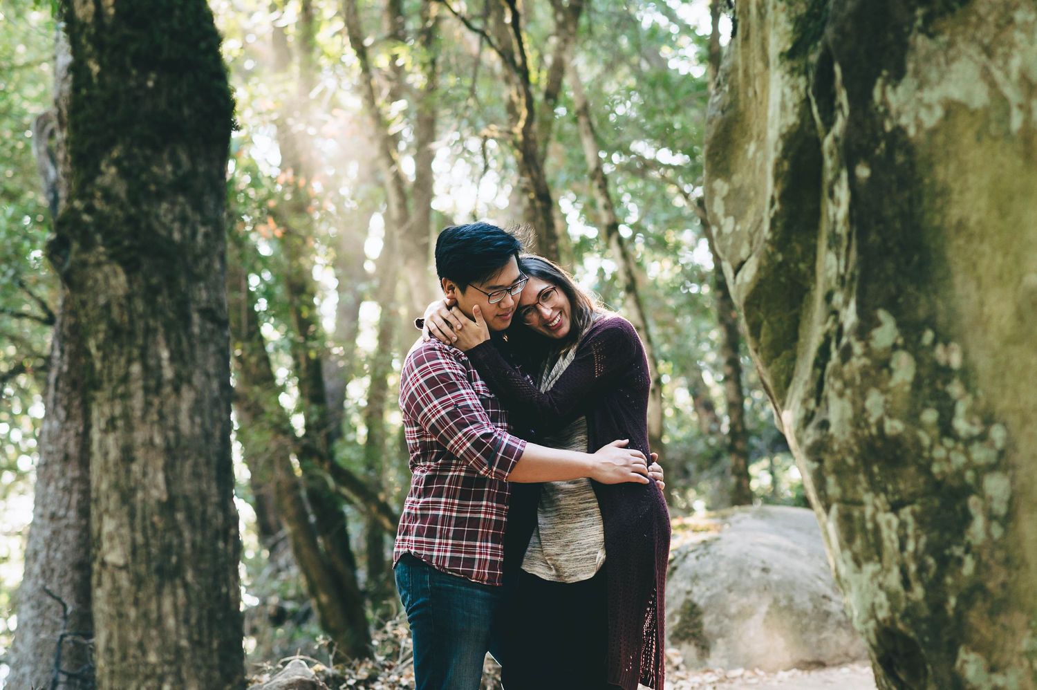 Two people embracing on a wooded path surrounded by tall trees.