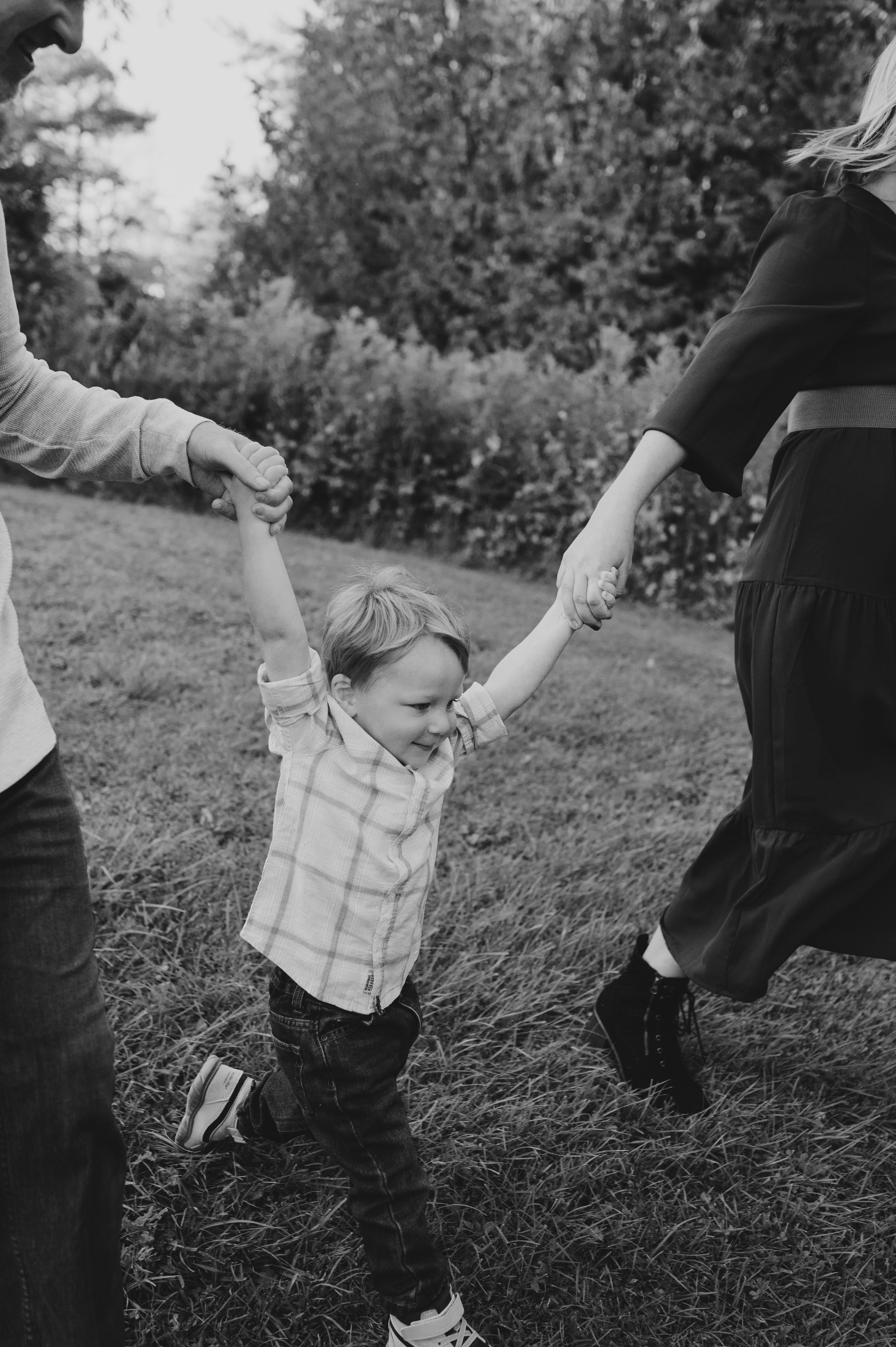 Black and white photo of a small child taking first steps while holding hands with adults.