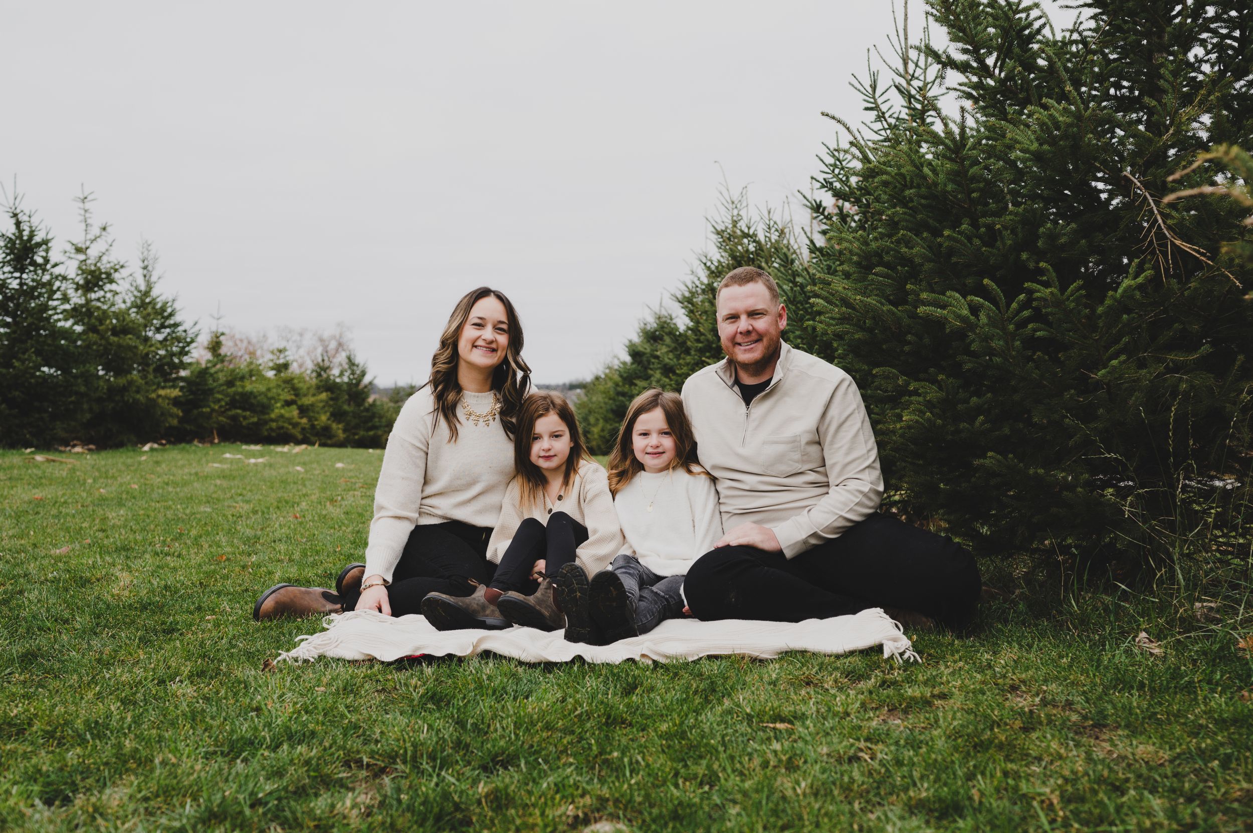 Family sitting together on a blanket in front of evergreen trees.