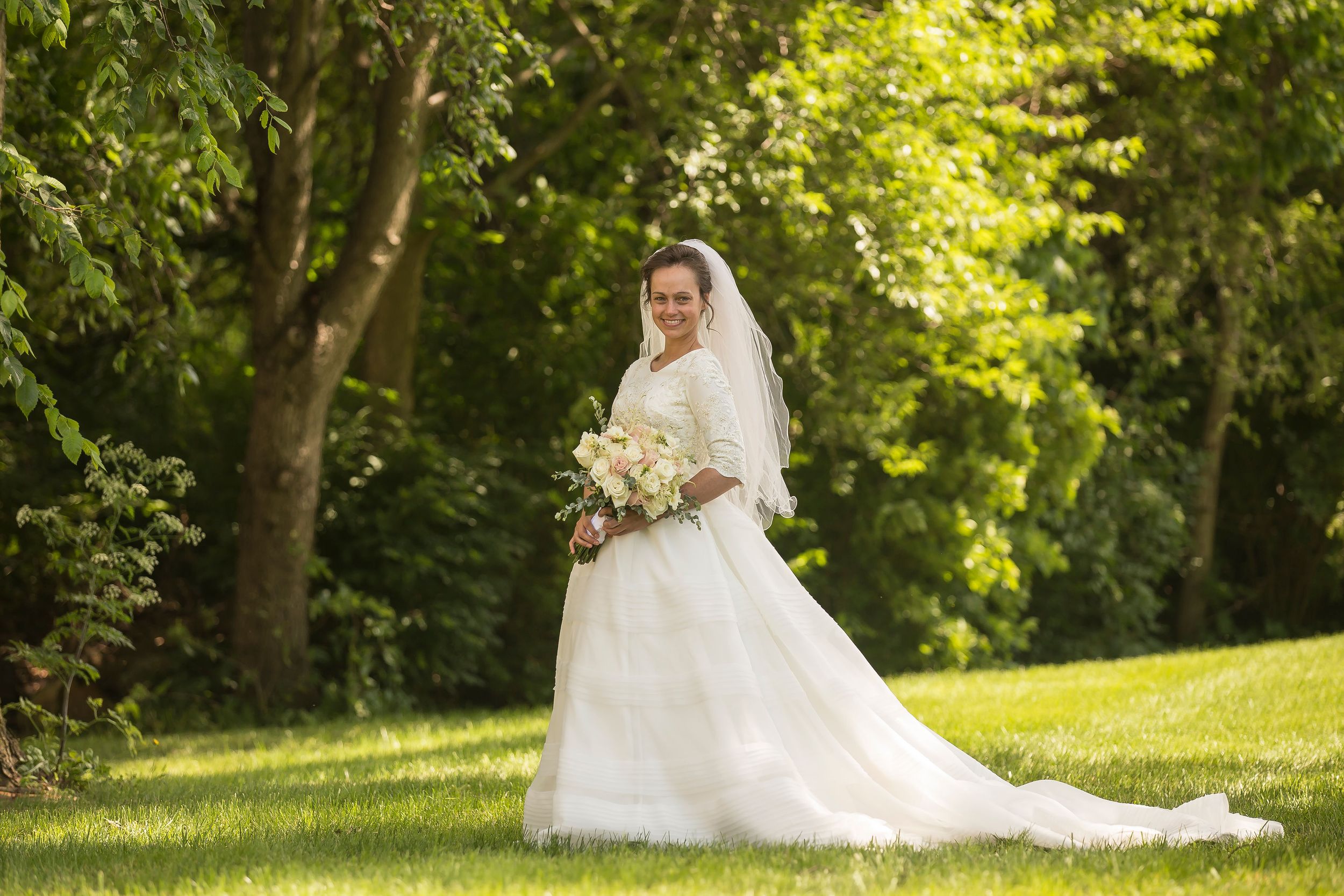 A bride in a white lace gown holds a bouquet while standing on a sunlit grassy hill.