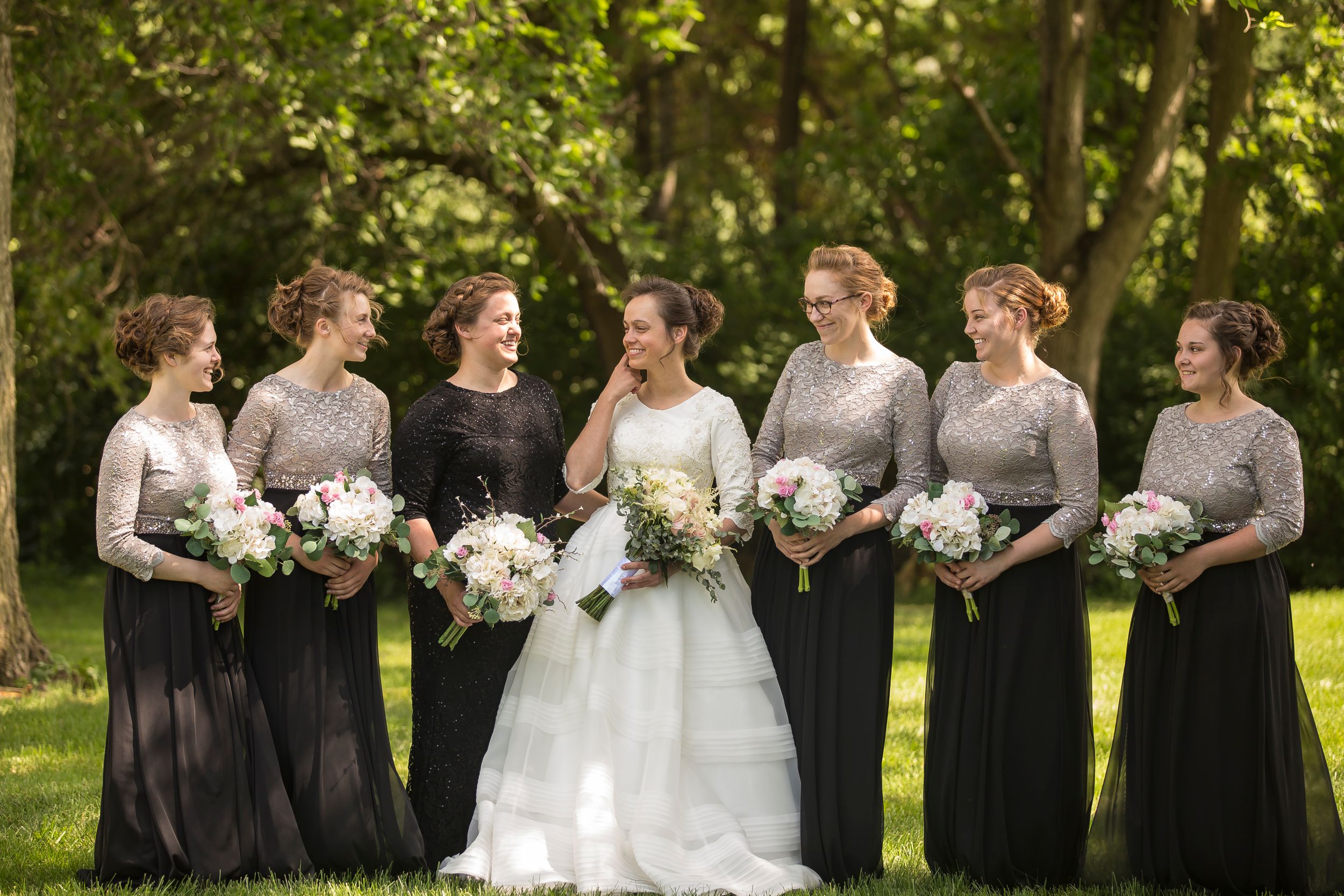 Wedding party wearing black and gray dresses poses outdoors in a lush green garden setting.