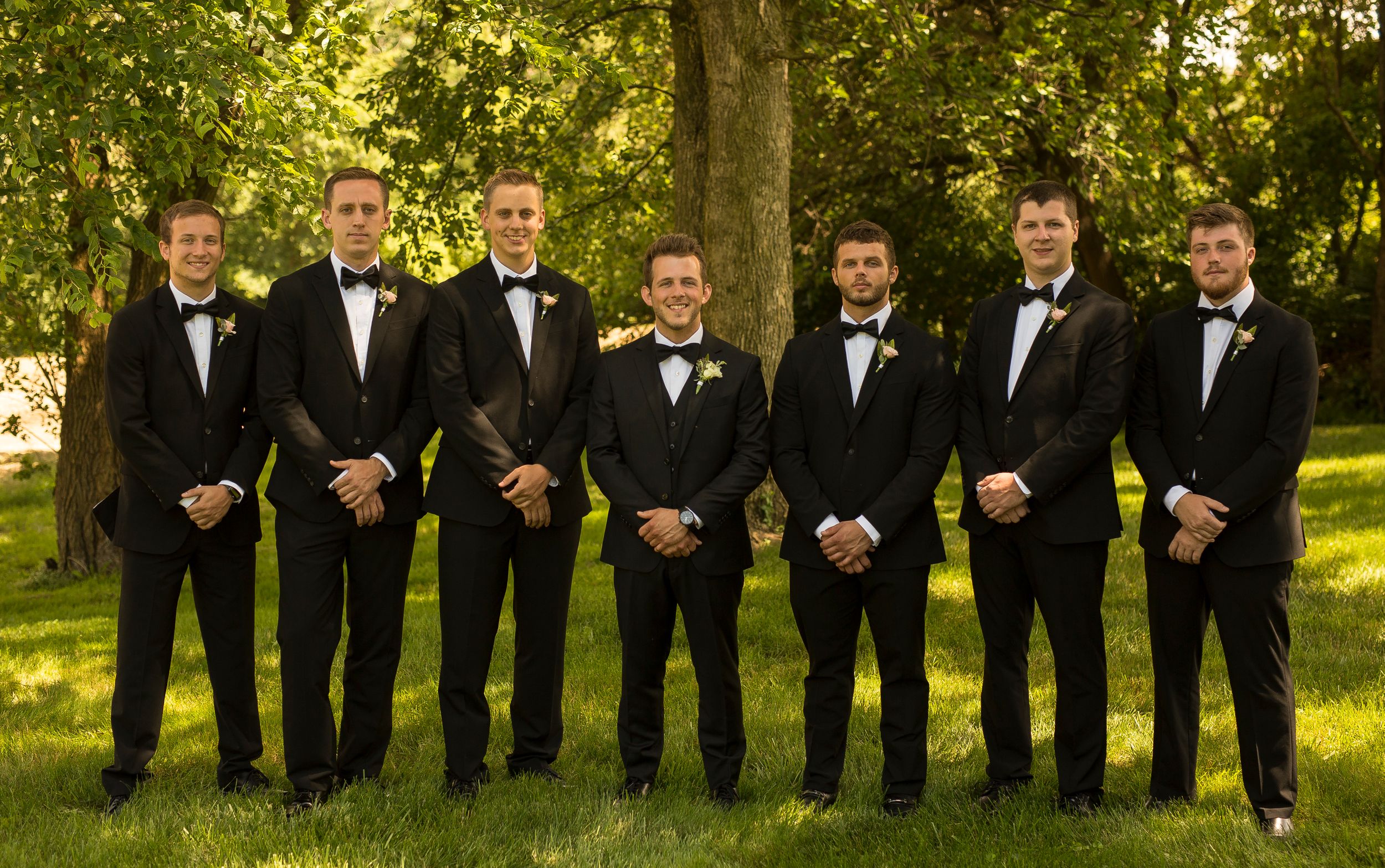 Groomsmen in black tuxedos lined up for formal wedding photo in garden setting.