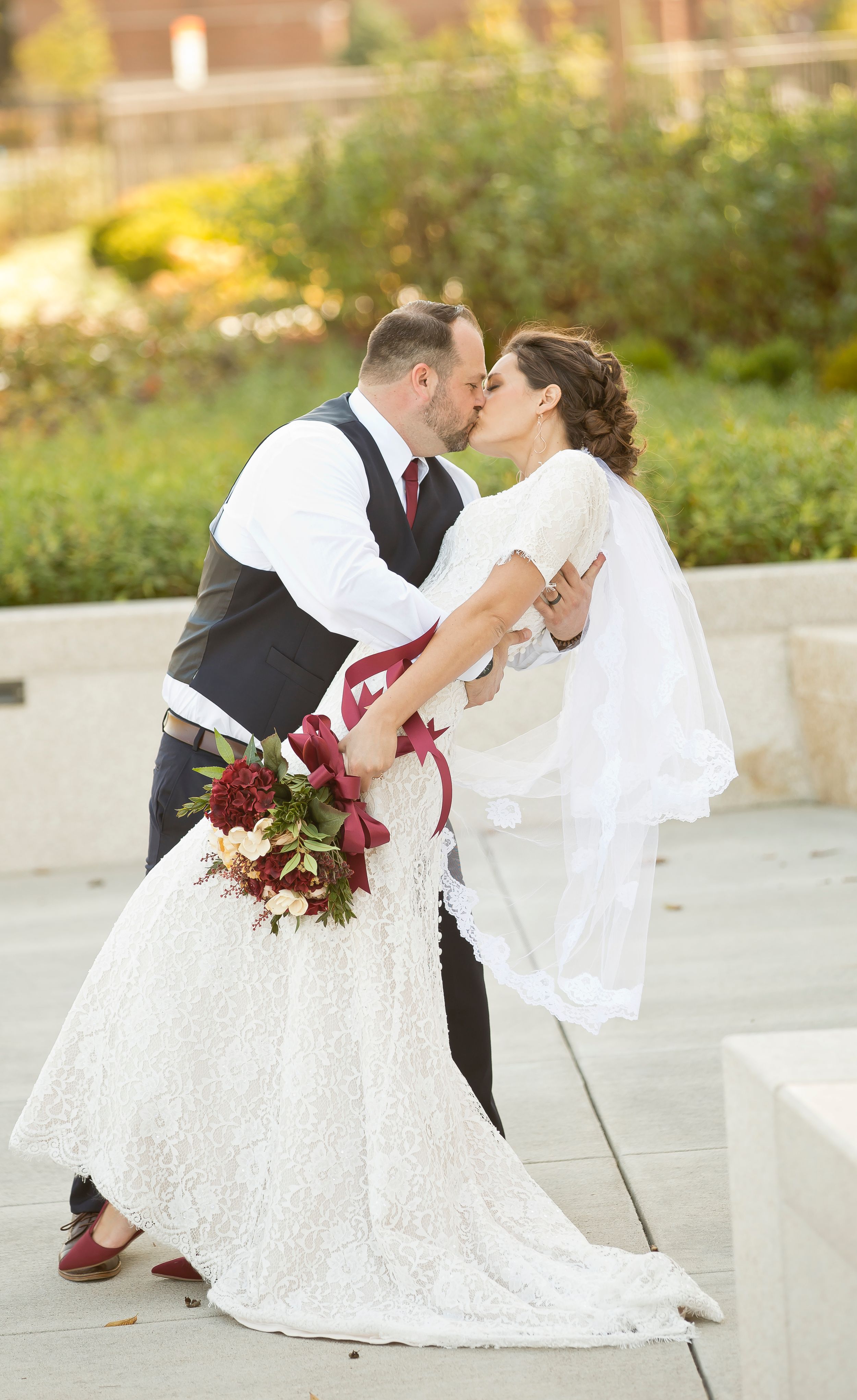 A romantic dip kiss moment captured with a burgundy bouquet in the foreground.