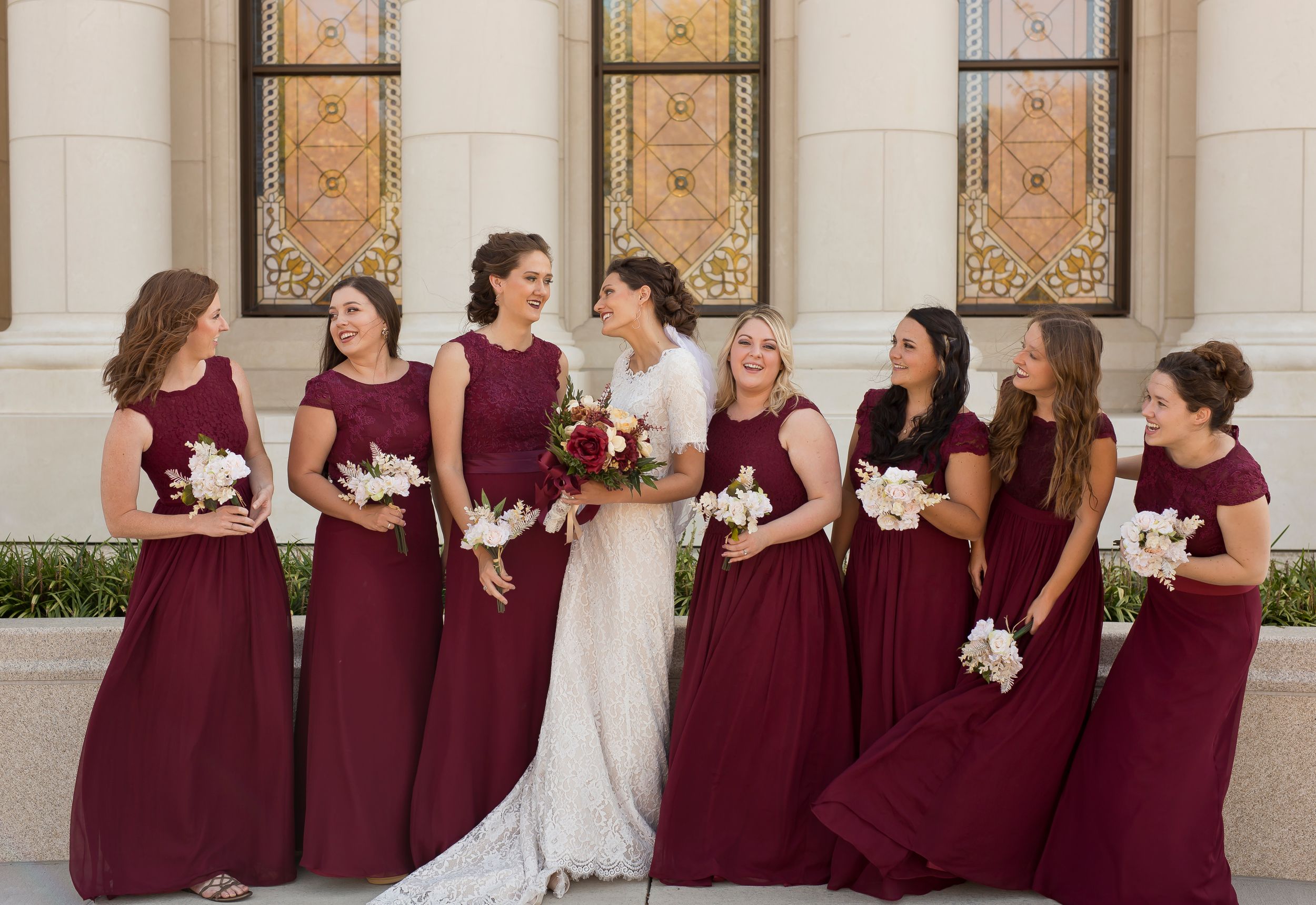 Bridesmaids in burgundy dresses stand together holding white bouquets.