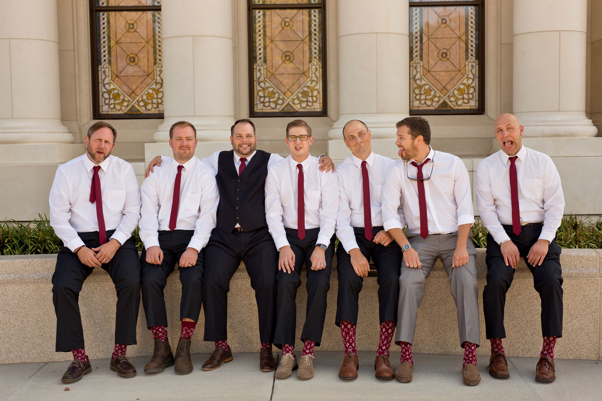 Groomsmen in white shirts and red ties pose casually against a stone building.