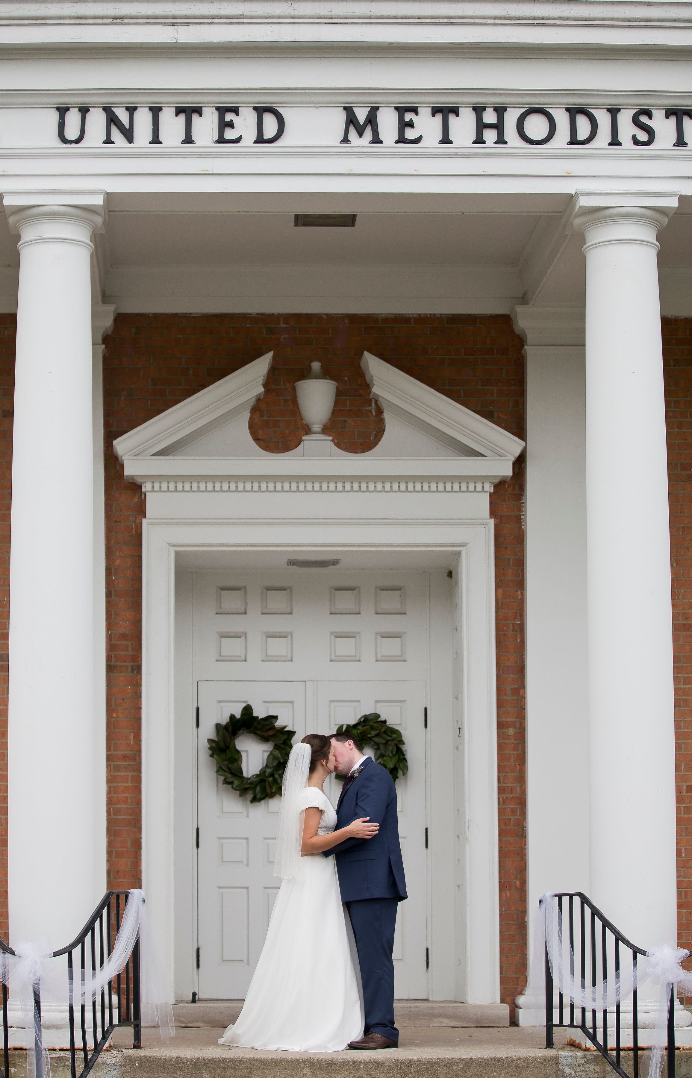 A wedding moment captured at the entrance of a United Methodist church with white columns.