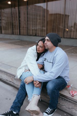couple sitting on a curb outdoors holding hands as one leans onto the other, sharing a quiet affectionate moment.