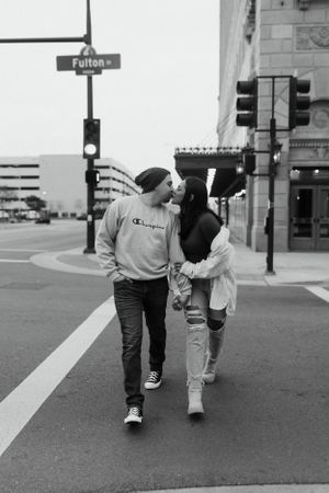 couple walking arm in arm across a city crosswalk, leaning in a for a kiss with traffic lights and buildings in the background