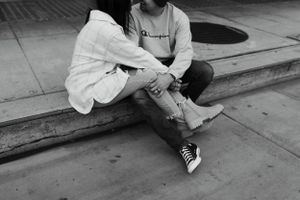 black and white photo of a couple sitting on a curb, holding hands with knees touching, leaning close in a quiet moment