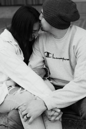 black and white photo of a couple sitting close on a curb outdoors, holding hands as one leans in for a gentle kiss on the forehead