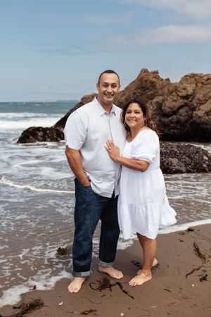 couple standing barefoot on a sandy beach, smiling at the camera as waves roll in behind them and rocky cliffs rise along the shoreline