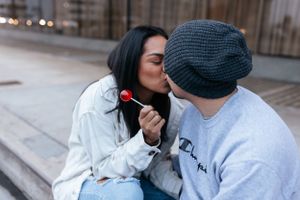 couple sitting close outdoors, sharing a kiss while one partner holds a red lolipop