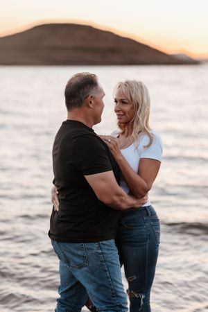 couple standing in shallow lake water at sunset, smiling and holding each other with hills in background