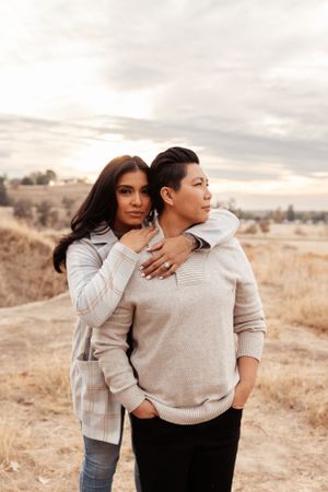 couple standing together in an open grassy landscape at golden hour, with one standing behind the other wrapping their arms around the other
