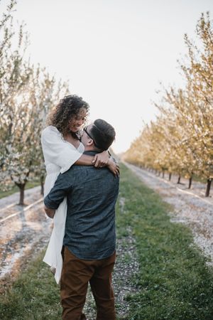 couple embracing in a almond orchard while in bloom, one partner is holding the other up by the waist and they are smiling at each other