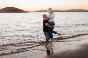 couple standing in shallow lake water at sunset, smiling and holding each other with hills in background