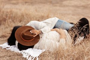 couple lying close together on a blanket in a golden field of tall grass, embracing softly as one covers their faces with a wide brim hat