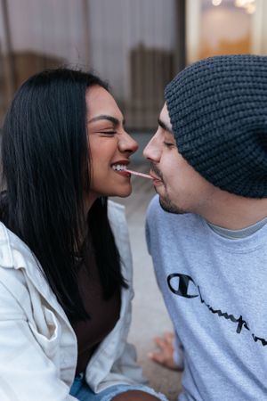 couple sitting close outdoors, smiling as they share a piece of candy between them, faces nearly touching in a playful moment