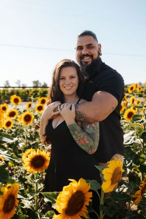 couple embracing in a sunflower field, smiling at the camera as bright yellow blooms surround them in midday sun