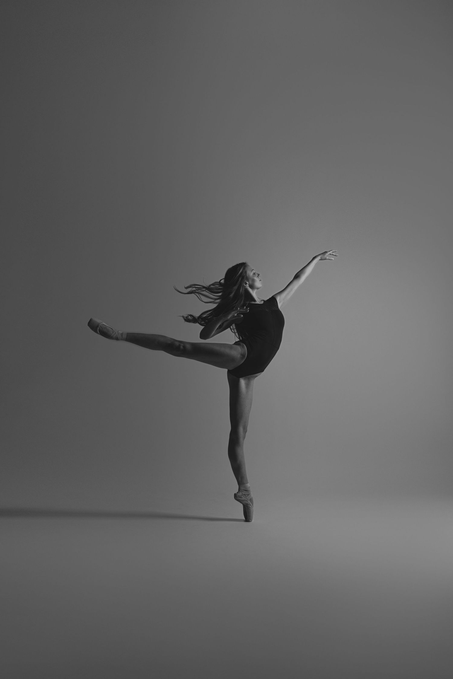 Graceful dancer in black performing arabesque pose in dramatic black and white studio lighting.