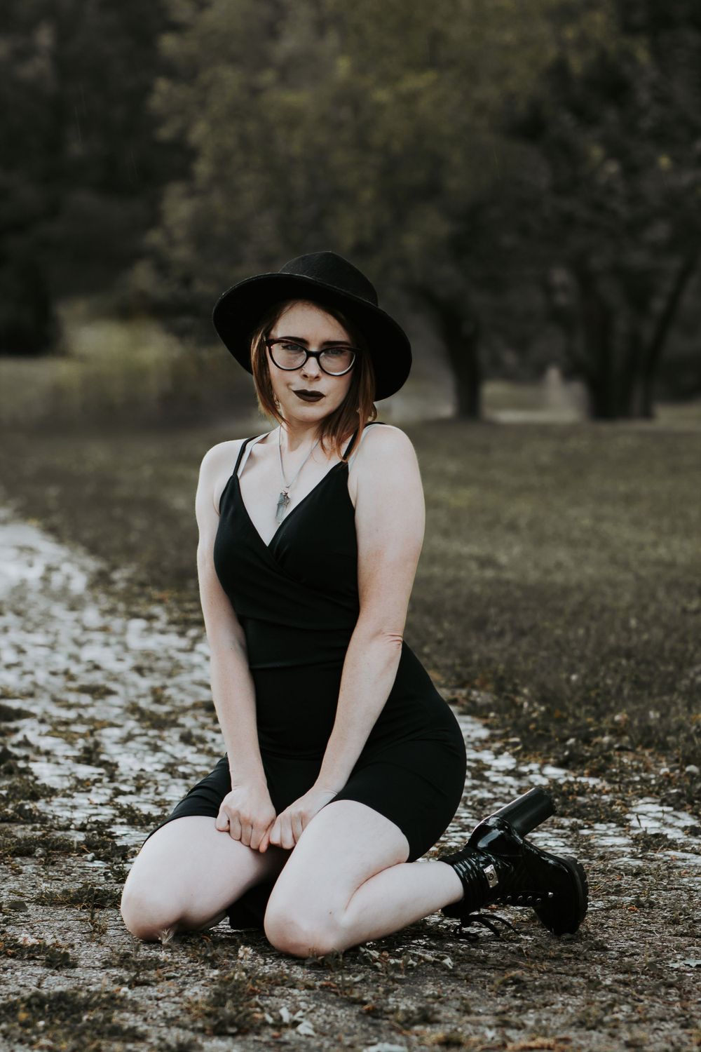 Person in black dress and hat sitting on ground with fallen leaves in moody outdoor setting.