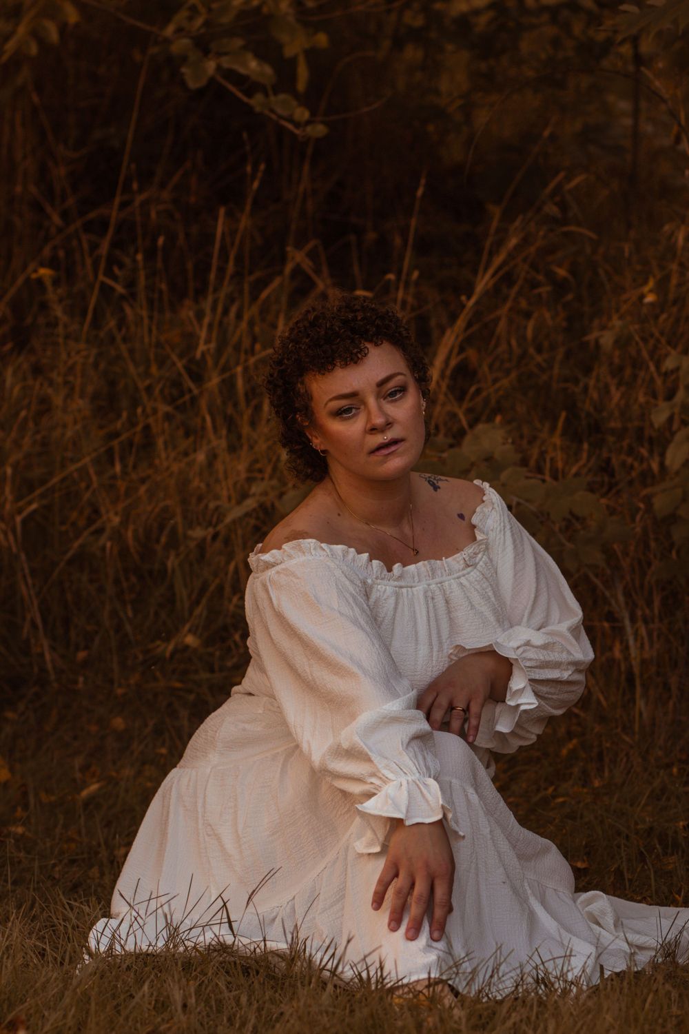 Person in white vintage dress sitting in field of dried grass at dusk.