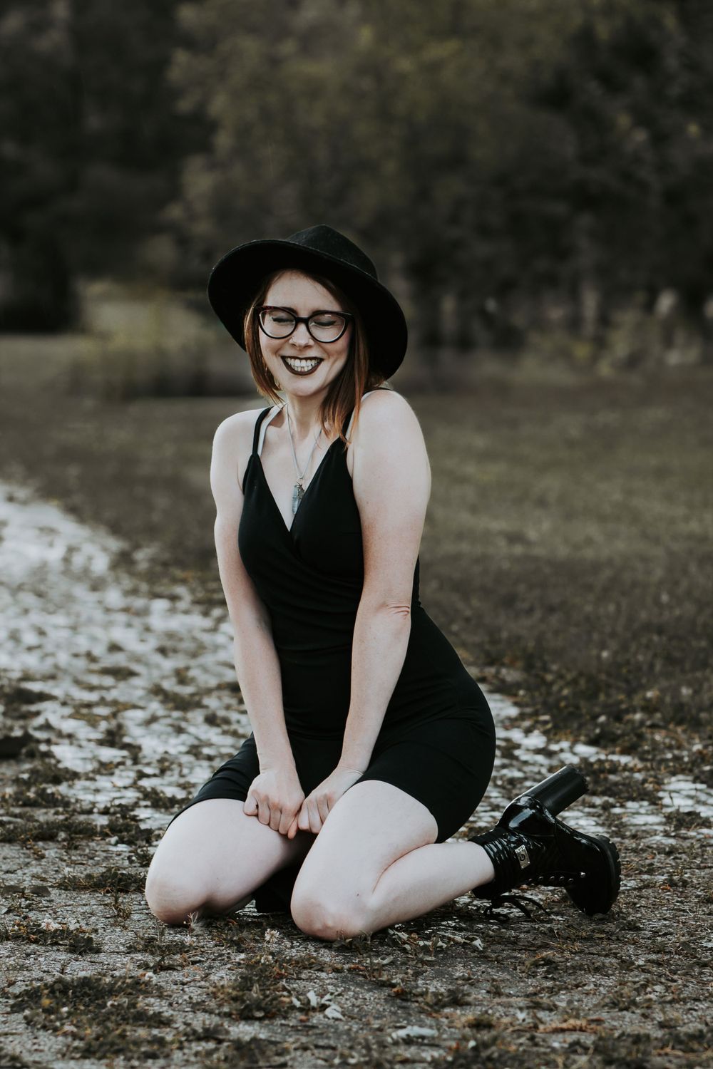 Figure in black dress and wide brim hat sitting among scattered leaves on ground.