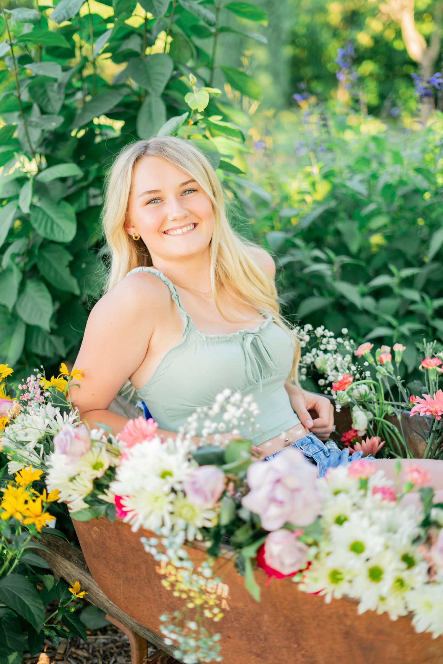 Smiling blonde woman in mint dress surrounded by colorful flowers in a lush garden setting.