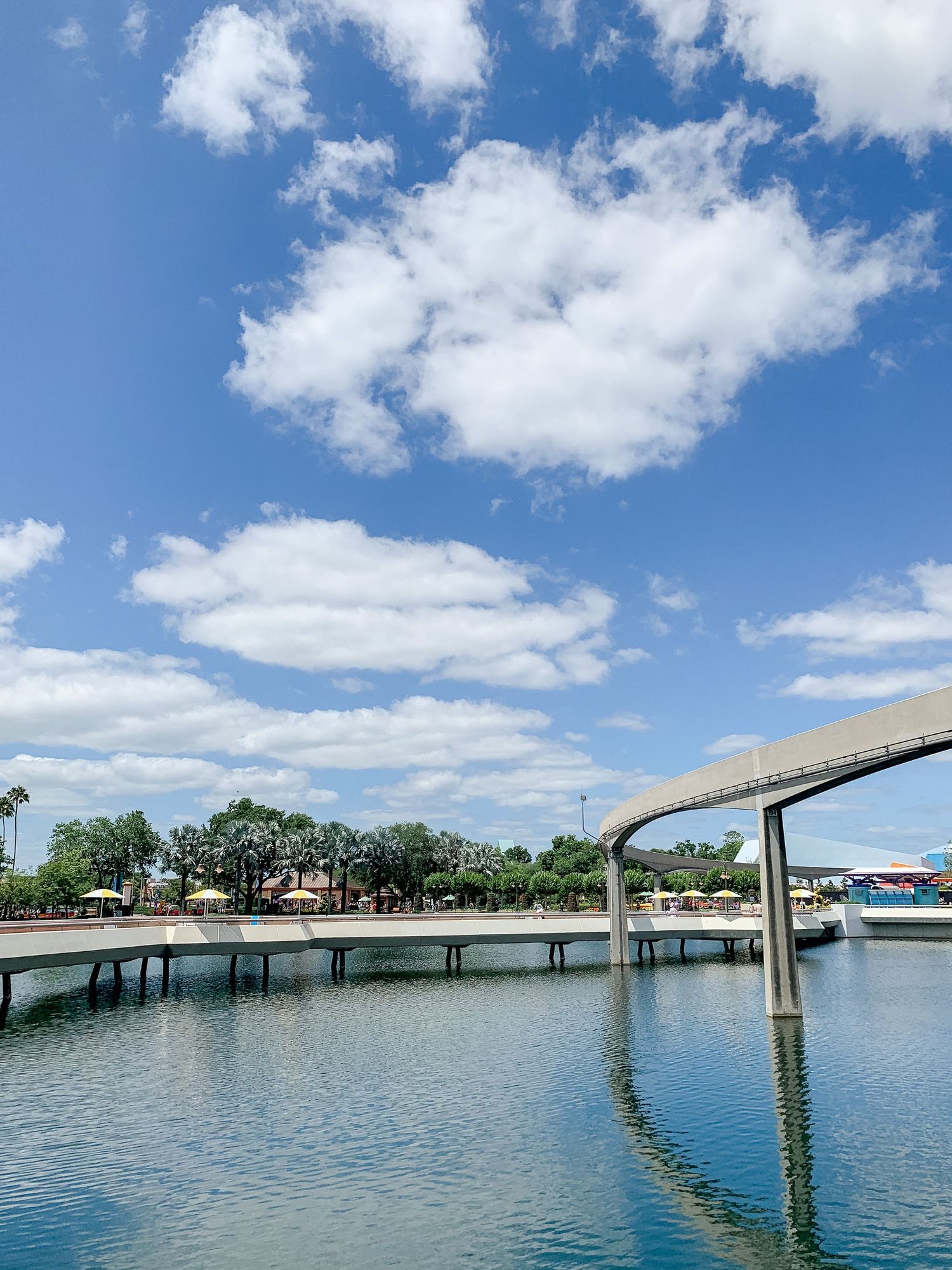 Monorail track curves over a tranquil lagoon with blue water and white clouds overhead.