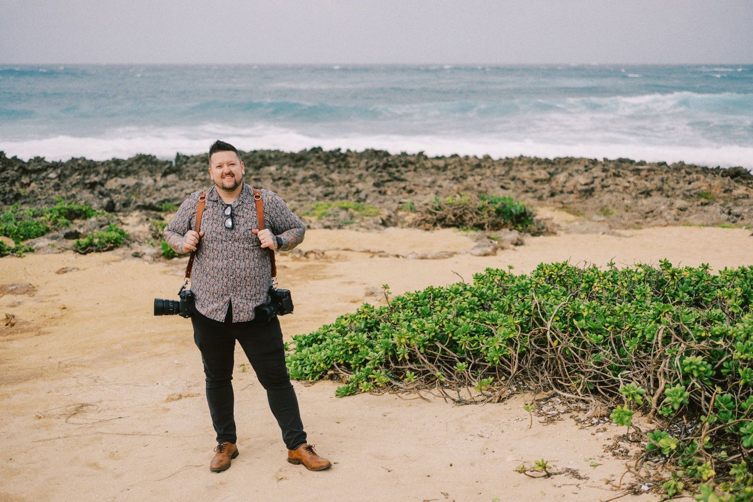 A photographer stands on a beach with camera gear against a tropical ocean backdrop.
