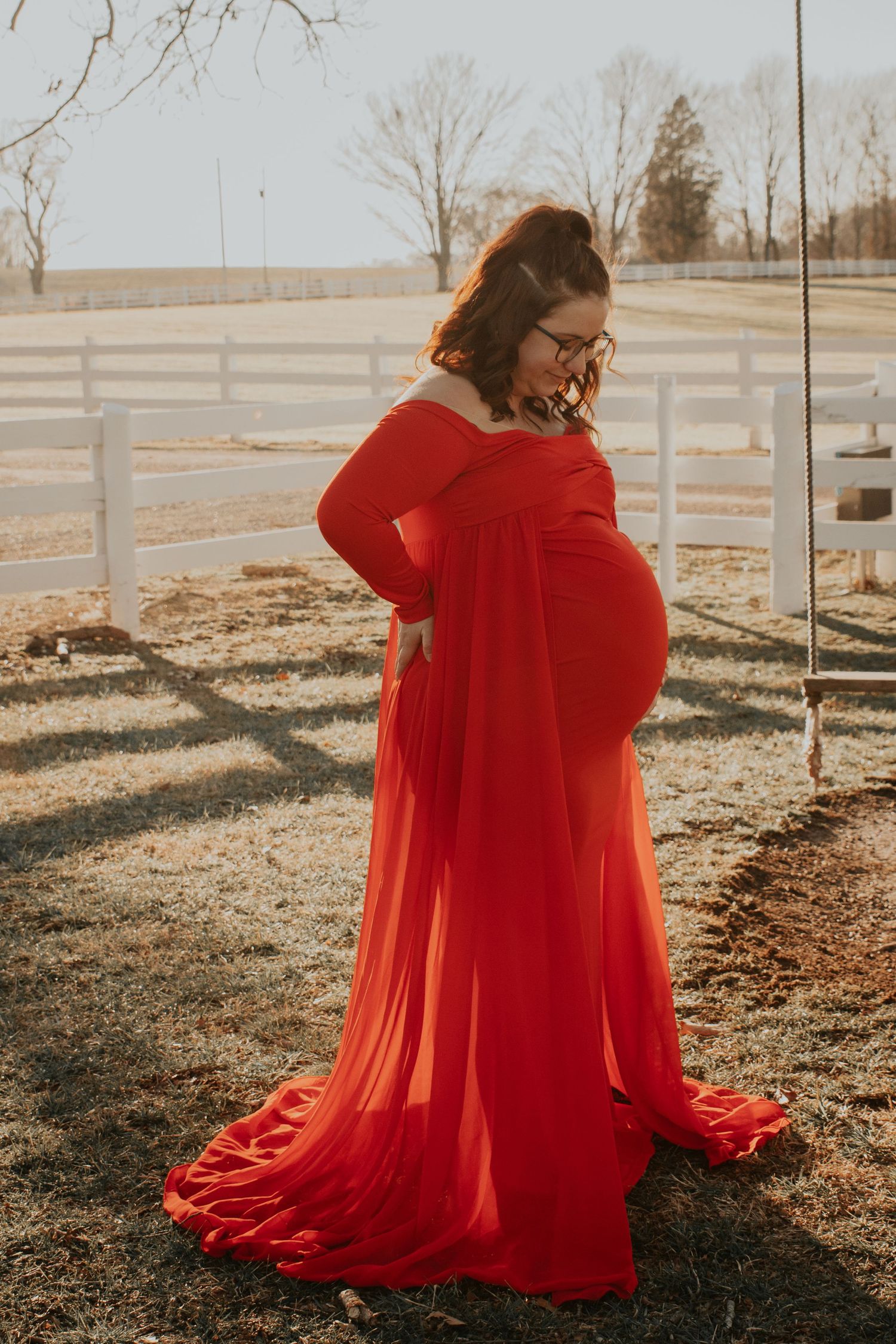 Woman in flowing red dress posing dramatically in sunlit field with white fence.
