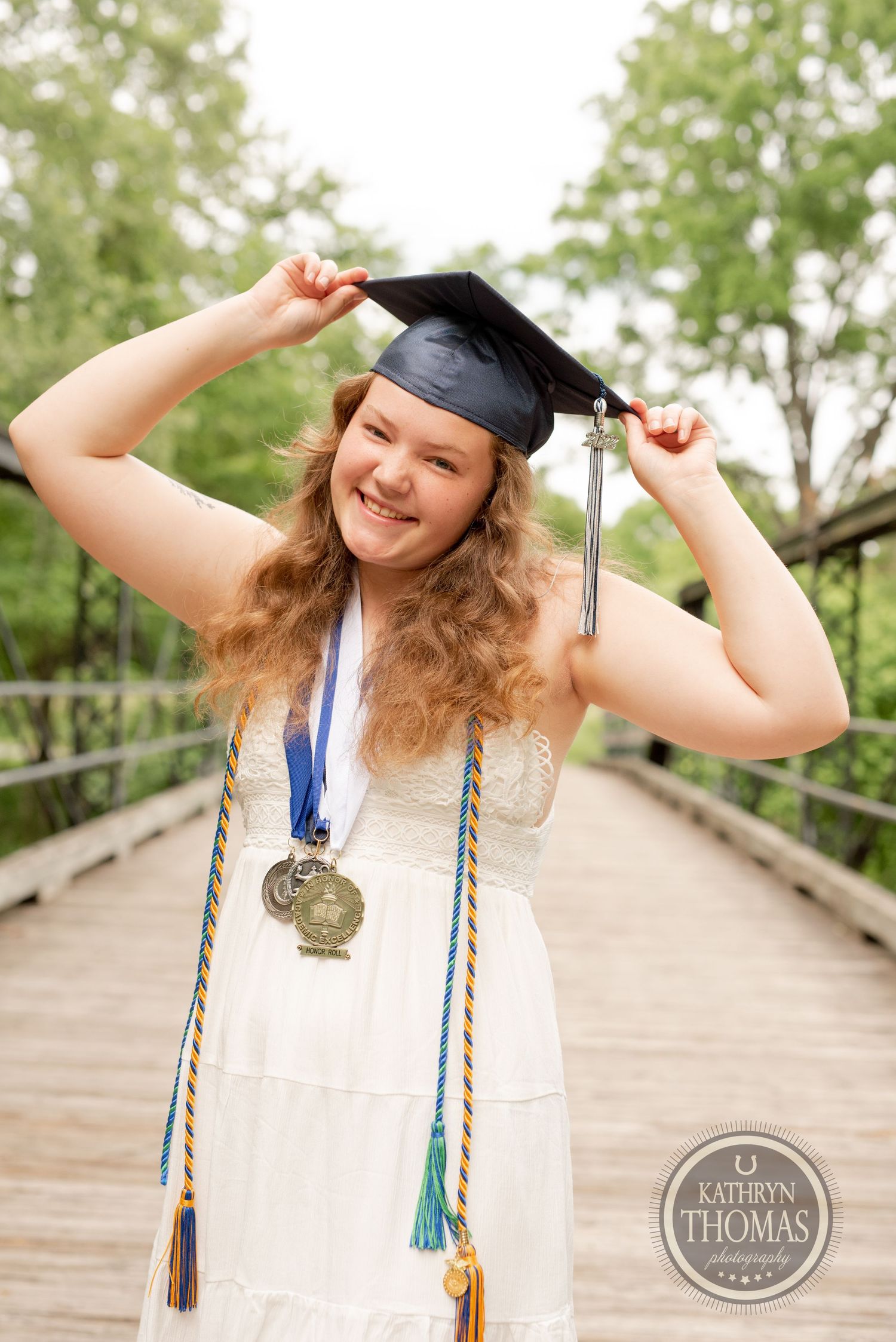 Graduate in white dress and cap holds tassel while standing on wooden bridge with academic regalia.