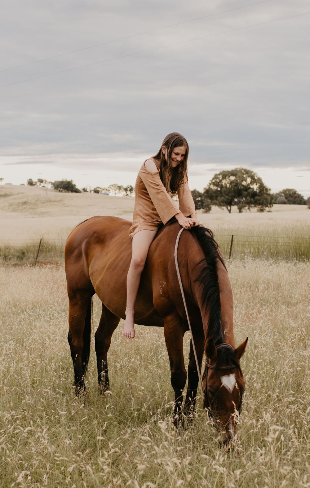 Girl bareback on her horse who is eating grass, standing in a paddock with a shed in the background
