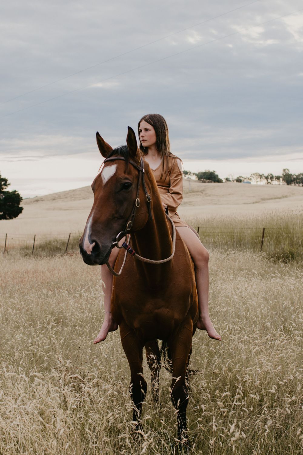 girl in caramel dress sitting bareback on her horse both looking the same way into the distance in a paddock with hills.
