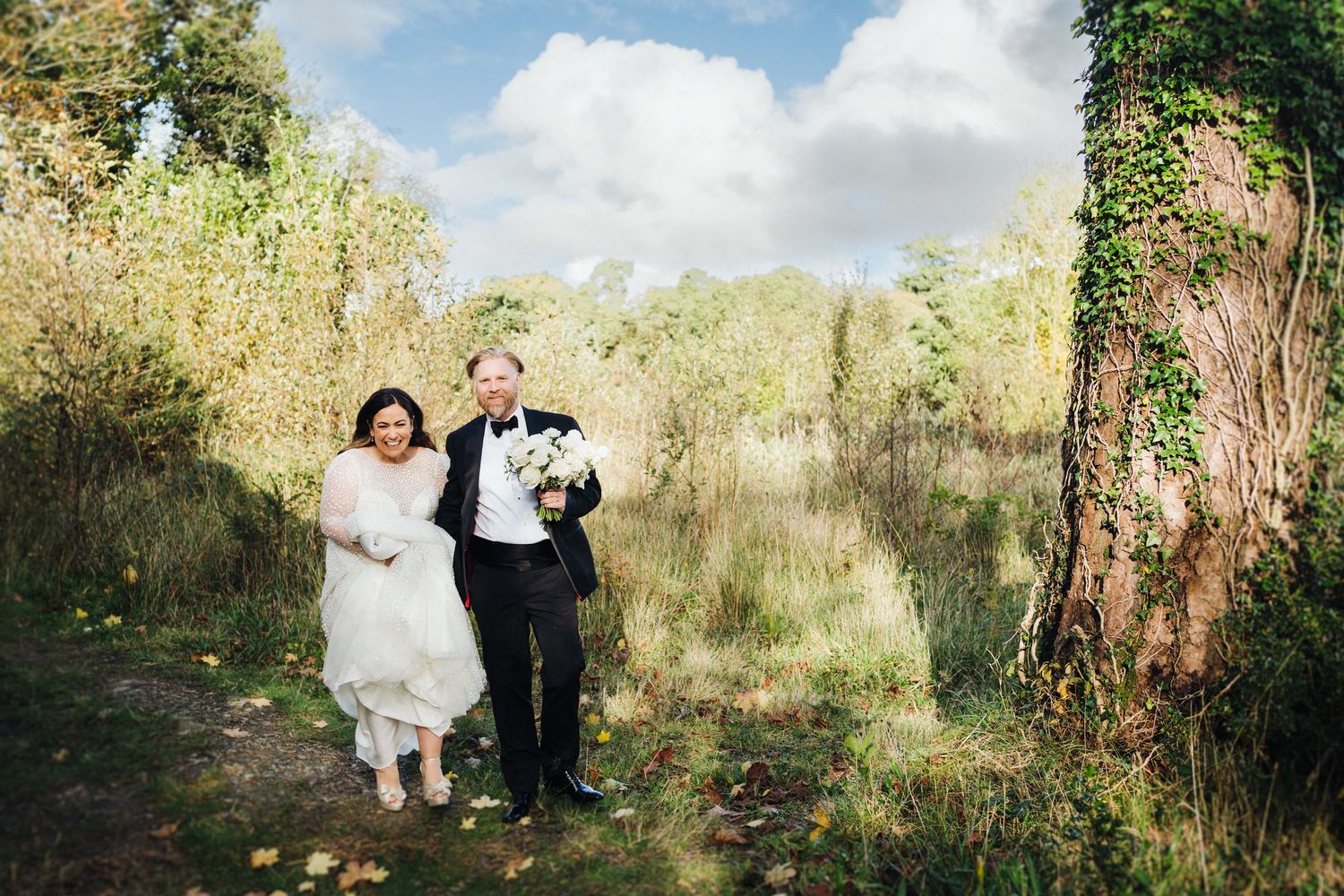Couple walks hand in hand through natural outdoor setting in formal wedding attire.