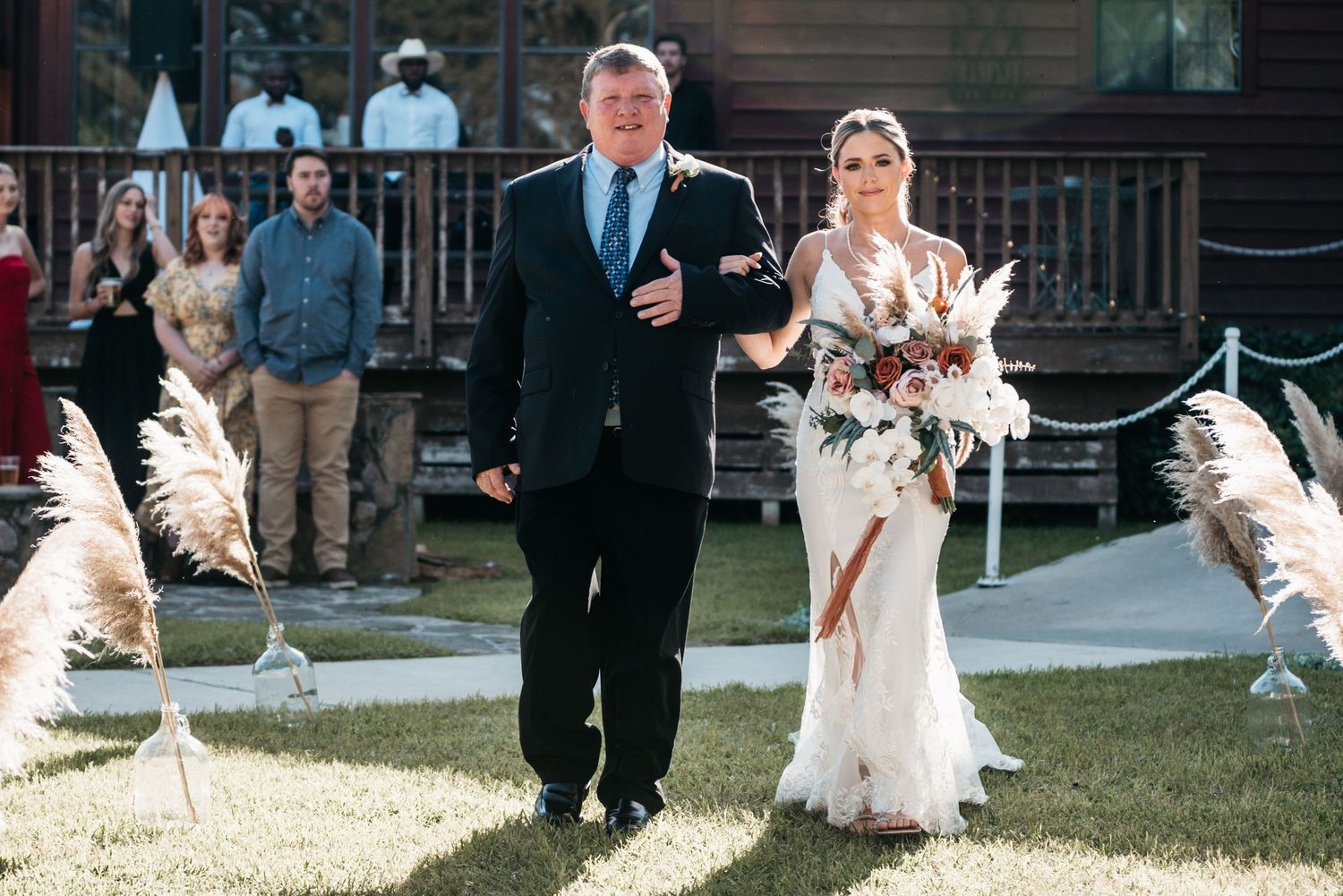 A wedding processional on a sunny day with pampas grass decorations lining the aisle.