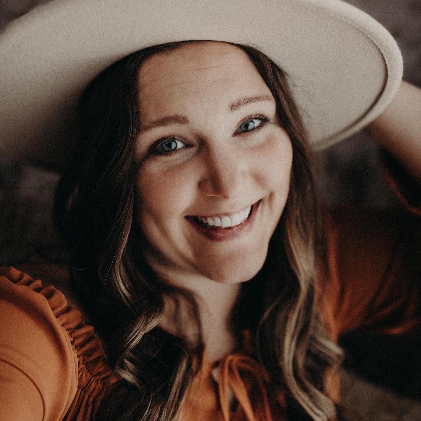 Portrait of smiling woman in wide brim hat and orange top with natural lighting.