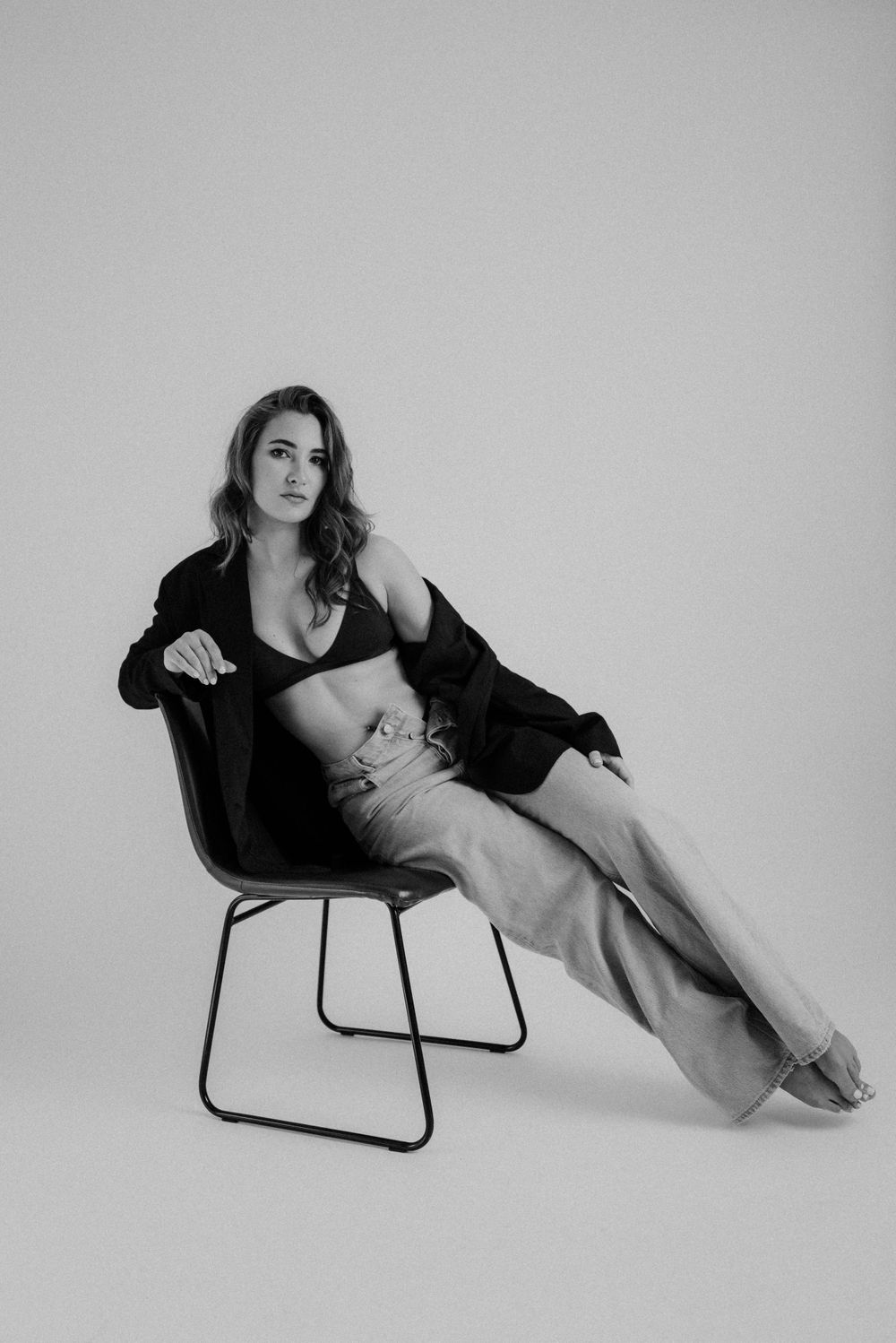 Woman in black outfit poses elegantly on modern chair during artistic photoshoot.