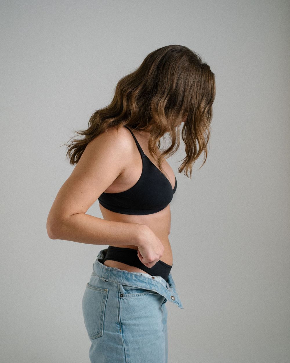 Side view of a woman wearing a black sports bra and jeans against a light background.